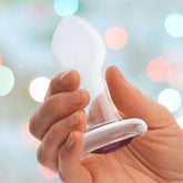 A hand holds a translucent object with a flat base and pointed tip, likely the Gläs 3.5" "Bling Bling" Glass Butt Plug. Crafted by Gläs, this product is ideal for prostate play and beginner-friendly. The blurred background features soft, colorful bokeh lights.